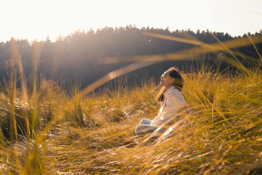 A woman sitting in a field of tall grass, looking up at the sun